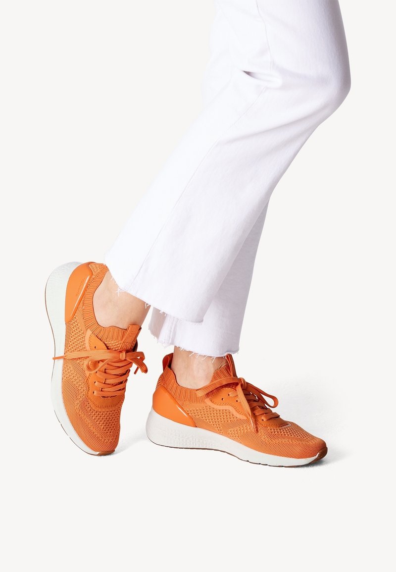 Orange Sport-Schuhe mit einem atmungsaktiven, gestrickten Obermaterial, strukturiertem Design und einer weißen Sohle, kombiniert mit weißen, cropped Hosen.