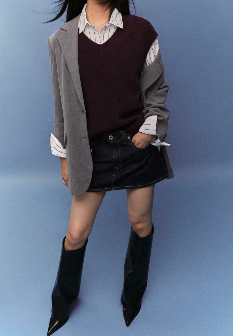 Femme portant un blazer gris sur une chemise rayée et un gilet bordeaux foncé, une mini-jupe en jean noire et des bottes noires montantes jusqu'aux genoux.
