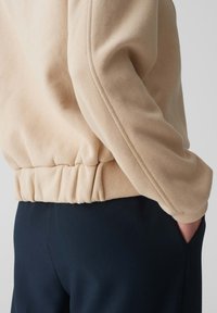 Felpa in pile beige con orlo arricciato e texture liscia, mostrata accanto a pantaloni blu scuro. Design semplice, con dettagli minimi.