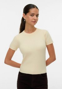 Maglione a maniche corte color crema lavorato a maglia con texture a costine e scollatura tonda, abbinato a pantaloni in velluto a coste marrone scuro.
