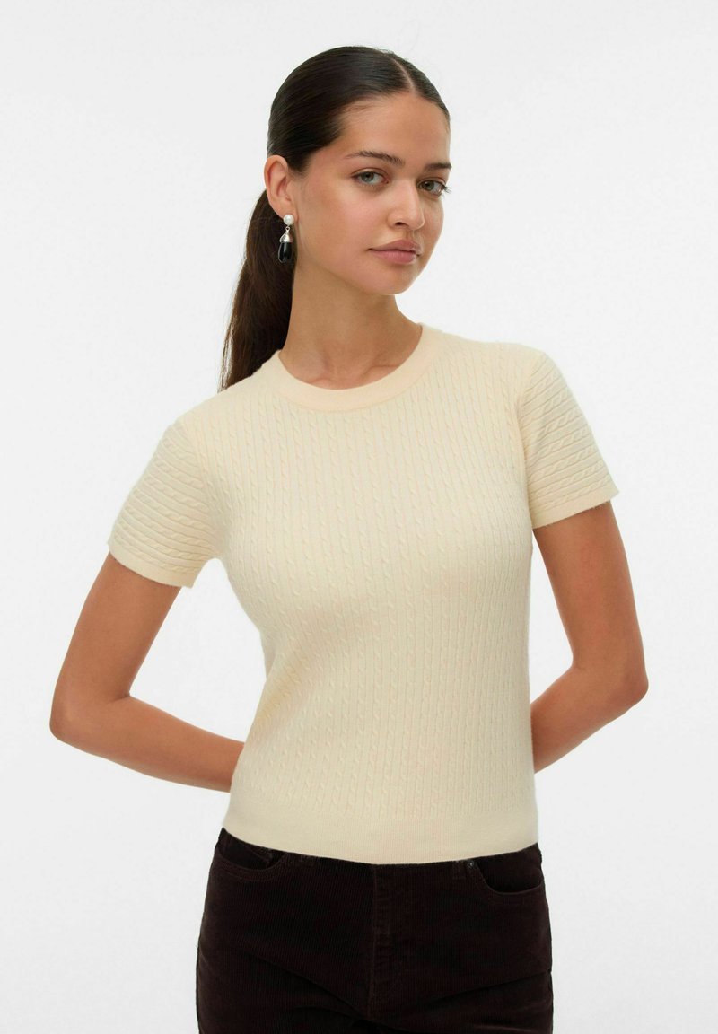 Maglione a maniche corte color crema lavorato a maglia con texture a costine e scollatura tonda, abbinato a pantaloni in velluto a coste marrone scuro.