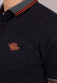 Laivastonsininen poolopaita, jossa on geometrinen kuvio, napillinen kaulus ja moniväriset raidalliset yksityiskohdat kauluksessa. Sisältää oranssin logotunnuksen.