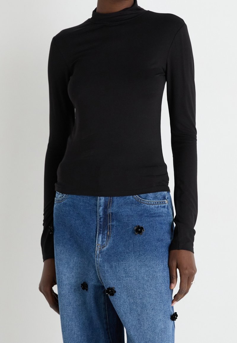 Haut à manches longues noir avec un col montant, fabriqué dans un matériau extensible, associé à un jean en denim bleu avec des accents floraux noirs.