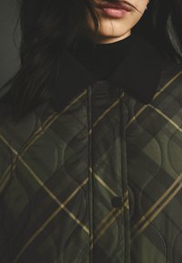 Jachetă verde quilted cu guler negru, cu un model de carouri aurii și verzi și detalii cu cusături mari, rotunjite.