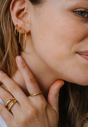 Frau mit drei goldenen Creolen in einem Ohr und drei goldenen Ringen an den Fingern, die ihren Hals berührt und leicht lächelt.