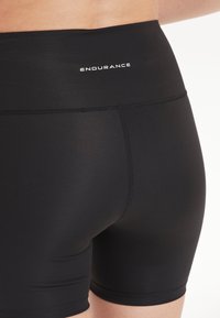 Pantaloni corti sportivi neri con un alto elastico in vita, dotati di testo "ENDURANCE". Realizzati in materiale liscio e elasticizzato con cuciture piatte.
