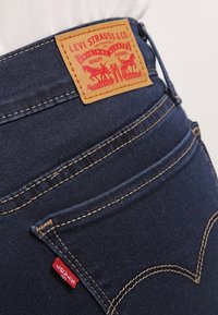 Jeans de mezclilla azul oscuro con un parche de cuero marrón claro que presenta gráficos rojos. Costura en un color contraste y una pequeña etiqueta roja visible.