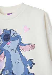 Sudadera crema con un personaje de dibujos animados azul, nariz brillante, orejas con manchas rosas y un diseño de corazón metálico rosa.
