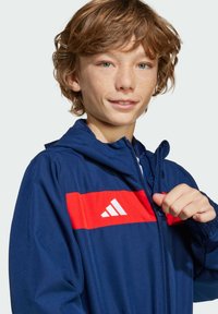 Veste marine avec une bande horizontale rouge et un logo blanc sur la poitrine, dotée d'une capuche et d'une fermeture éclair, fabriquée en tissu lisse.