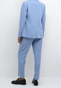 Osoba ubrana w jasnoniebieski, dopasowany garnitur z prostymi spodniami i czarnymi skórzanymi mokasynami, stojąca na tle gładkiej białej ściany.