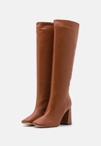 Bottes en cuir marron montantes avec un talon carré, présentant une texture lisse et un design à bout carré. Silhouette classique et épurée.