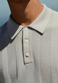 Lichtgrijze poloshirt met een gestructureerd breisel, voorzien van een klassieke kraag en vier witte knopen aan de voorkant.
