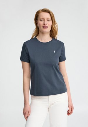 Woman with light skin and red hair wearing a dark gray short-sleeve t-shirt with a small white logo, paired with white pants, against a plain background.