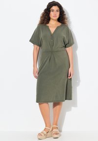 Oliwkowa sukienka do kolan z krótkimi rękawami, dekoltem w kształcie litery V, zapięciem na guziki i wiązanym pasem. Połączona z beżowymi sandałami na platformie.