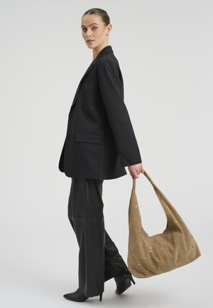 Ženska v črnem suknjiču in hlačah drži veliko svetlo rjavo semišasto torbo z zlatimi okraski, stoji pred enostavnim svetlim ozadjem.