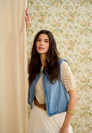 Kvinde med langt bølget hår iført en beige striktrøje, blå vest med flettede kanter, hvid nederdel og gyldt armbånd, stående ved en gardin.