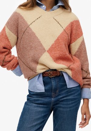 Personne portant un pull douillet à motif losanges en beige, rouille et rose, par-dessus une chemise à col bleue, avec un jean bleu et une ceinture tressée marron.