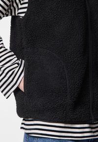 Crni prsluk od flisa s teksturiranim tkanjem, zatvaračem na patentny zatvarač i bočnim džepovima. Nošen preko prugaste majice dugih rukava s crno-bijelim prugama.