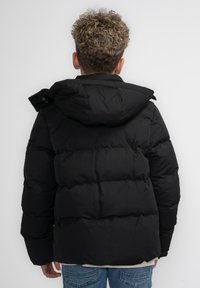 Giacca a vento nera con cappuccio, caratterizzata da un design trapuntato, tessuto testurizzato e una vestibilità rilassata. Camicia di colore chiaro e jeans in denim visibili.