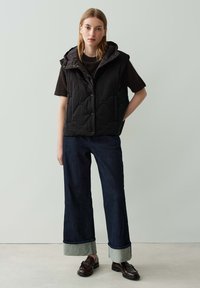 Schwarze gesteppte Weste mit Kapuze, ausgestattet mit Druckknöpfen. Kombiniert mit weiten, dunklen Jeans mit umgeschlagenen Säumen und braunen Slippern.