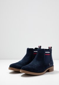 Botas de tobillo de ante azul marino con paneles laterales elásticos que presentan rayas rojas y blancas. Suela de goma marrón, punta redonda y tacón bajo.