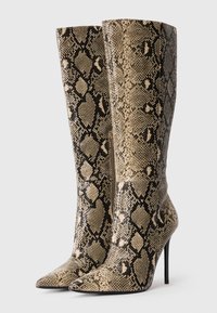 Un paio di stivali alti fino al ginocchio con tacco a stiletto e punta affusolata, con un motivo in pelle di serpente beige-nera, posizionati uno accanto all'altro su uno sfondo bianco.