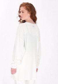 Dlouhý bílý top z lehké tkaniny, s volným střihem, kulatým výstřihem a mírně oversized rukávy s nařasenými manžetami.