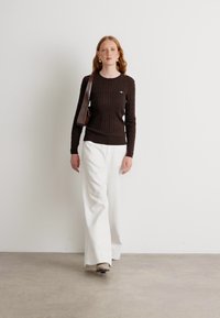 Pull en tricot marron avec un ourlet côtelé, associé à un pantalon large blanc. Les accessoires comprennent un sac à bandoulière marron et des boucles d'oreilles.
