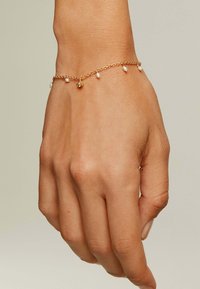 Pulsera de oro con pequeñas perlas blancas y un charm decorativo, con delicados eslabones de cadena y un acabado pulido, llevada en una mano.