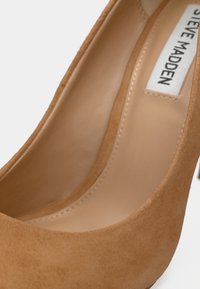 Κοντινό πλάνο εσωτερικού τακουνιού από καφέ σουέτ δέρμα με ραμμένη σόλα και ετικέτα με την επωνυμία Steve Madden.