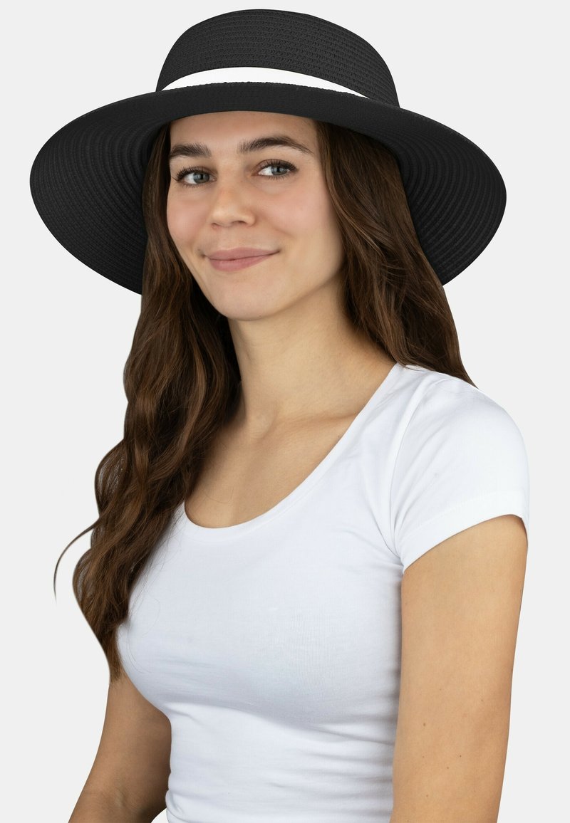 Schwarzer, breitkrempiger Hut mit strukturierten Oberflächen, versehen mit einem weißen Band. Getragen von einem Model mit langen braunen Haaren vor einem neutralen Hintergrund.