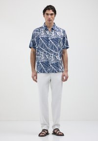 Uomo che indossa una camicia a maniche corte blu e bianca a fantasia, pantaloni bianchi e sandali neri, in piedi davanti a uno sfondo bianco semplice.