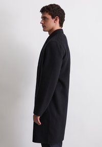 Ein Mann steht seitlich und trägt einen langen schwarzen Wollmantel mit Kragen vor einem schlichten weißen Hintergrund.