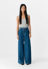 Lichtgrijze mouwloze top gecombineerd met hoge taille, wijde, donkerblauwe denim jeans. De jeans hebben een strikkoord in de taille.