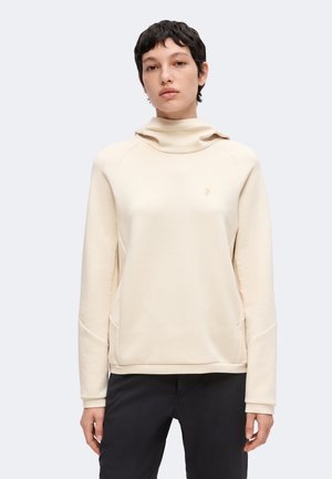 Personne aux cheveux courts et foncés portant un haut crème à col montant et manches longues et un pantalon noir, sur un fond uni clair.