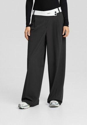 Pantalon noir à jambes larges avec une ceinture blanche asymétrique maintenue par deux boutons noirs, porté avec des baskets blanches et un top noir à manches longues.