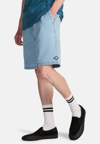 Jasnoniebieskie sztruksowe szorty z pionowymi paskami, bocznymi kieszeniami i czarną naszywką z logo, w zestawie z czarnymi butami wsuwanymi i białymi skarpetkami w paski.