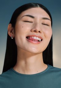 Primo piano di una persona con capelli neri e lisci, che indossa una camicia turchese. Hanno gli occhi chiusi, un leggero sorriso e una lingua visibile.