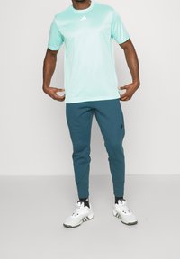 Man i ljusblå kortärmad t-shirt, mörkblå joggerbyxor, svarta strumpor och vita Adidas sneakers, stående mot vit bakgrund.