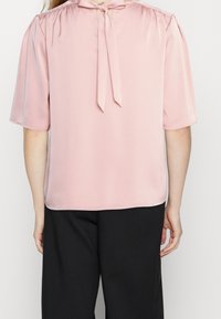 Blusa rosa chiaro realizzata in tessuto morbido, con maniche corte, un nodo al collo e un taglio rilassato. Indossata con pantaloni neri.
