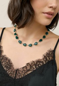 Smaragdgrünes Schmuckhalsband mit goldfarbener Beschichtung, das unregelmäßige Formen und verbundene Fassungen aufweist. Getragen über einem schwarzen Spitzen-Camisole.