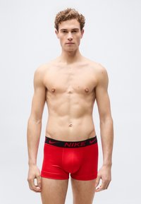 Νέος άνδρας με σγουρά μαλλιά στέκεται γυμνόστηθος φορώντας κόκκινα ανδρικά εσώρουχα Nike με μαύρη μέση, μπροστά σε απλό ανοιχτό γκρι φόντο.