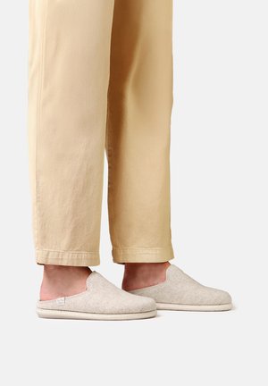 Pieds portant des chaussons beiges à enfiler avec dessus en textile et semelles rembourrées, associés à un pantalon beige clair à jambes droites.
