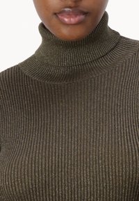 Gros plan sur une personne portant un pull col roulé métallique côtelé vert olive, montrant uniquement le cou et les lèvres.