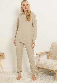 Donna in piedi a piedi nudi che indossa un completo loungewear beige a costine con colletto zip, accanto a una sedia di legno imbottita beige e a un piccolo tavolo di legno.