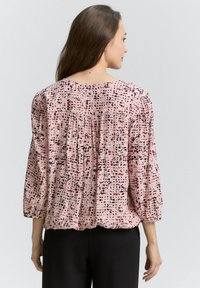 Bluză roz cu model, cu mâneci scurte tip balon, dintr-un material texturat și cu un detaliu la spate, purtată peste pantaloni închisi la culoare.