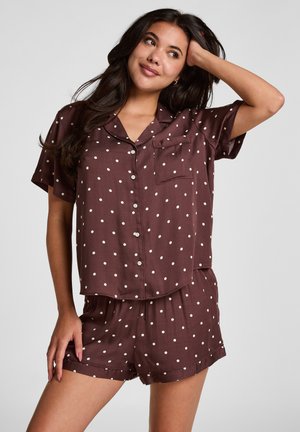 Femme portant une chemise marron à manches courtes avec boutons et un short assorti à pois blancs, posant avec une main sur la tête.