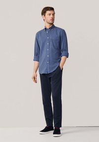Camisa azul de botones con mangas enrolladas, combinada con pantalones oscuros y zapatillas negras. Tiene un bolsillo en el pecho y una tela texturizada.