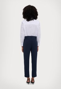 Chemise blanche à boutons avec des détails texturés sur les manches, assortie à un pantalon bleu marine ajusté et des sandales à talons noires, vue de dos.