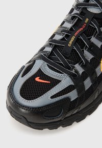 Baskets noires et grises avec des matériaux en maille et en cuir, présentant des accents orange et jaune, un laçage complexe et une semelle texturée.